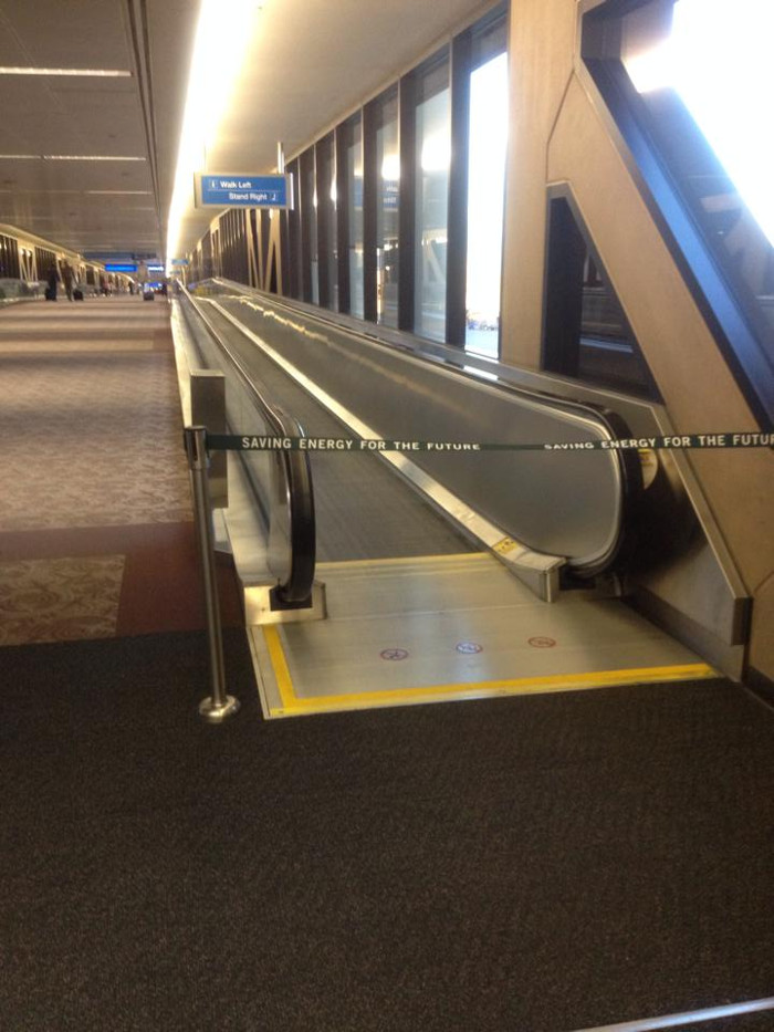 The photograph
depicts a long carpeted airport hallway with a moving walkway that
takes up the right half of the image. The walkway seems to still be
in operation, but it is blocked with a barricade labeled &ldquo;Saving
Energy for the Future.&rdquo;
