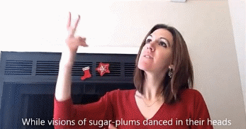Figure 2 shows me, a young woman with shoulder-length brown hair, wearing a red long-sleeve shirt with stockings on a chimney behind me. I am signing VISIONS and looking upward while white captions at the bottom of the screen read &ldquo;While visions of sugar-plums danced in their heads.&rdquo;