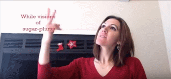 Figure 3 shows me, a young woman with shoulder-length brown hair, wearing a red long-sleeve shirt with stockings on a chimney behind me. I am signing VISIONS and looking upward towards red captions that appear in the upper left corner. My eyes and signs are directed towards the space that the captions appear in. The captions read &ldquo;While visions of sugar-plums.&rdquo;