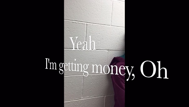 This screenshot shows a white wall and part of a woman's shoulder leaning on the wall. Large white captions appear diagonally on the screen and read &ldquo;Yeah I'm getting money, Oh.&rdquo;